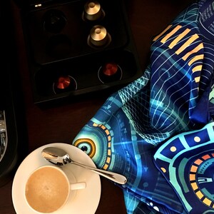 Puede incluir: Una taza de caf&eacute; con una cuchara sobre un platillo blanco, junto a una m&aacute;quina de espresso negra y una bufanda azul estampada. La bufanda tiene un dise&ntilde;o geom&eacute;trico con detalles turquesa, amarillos y blancos. C&aacute;psulas de caf&eacute; en un soporte negro.