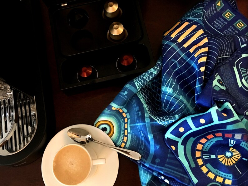 Puede incluir: Una taza de caf&eacute; con una cuchara sobre un platillo blanco, junto a una m&aacute;quina de espresso negra y una bufanda azul estampada. La bufanda tiene un dise&ntilde;o geom&eacute;trico con detalles turquesa, amarillos y blancos. C&aacute;psulas de caf&eacute; en un soporte negro.