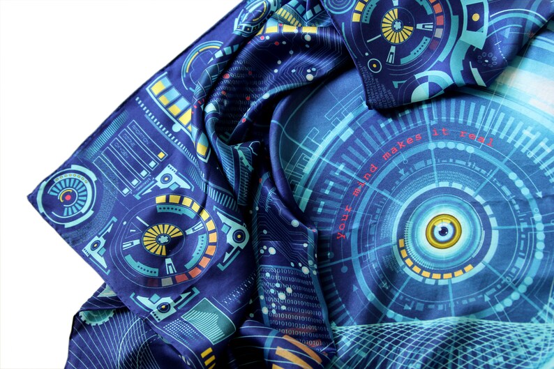 Puede incluir: Bufanda azul oscuro con un dise&ntilde;o futurista con patrones de circuitos y gr&aacute;ficos circulares en tonos turquesa, amarillo y naranja. El texto "Your mind makes it real" es visible. La bufanda est&aacute; doblada y drapeada.