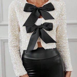 May include: A white sequined cropped jacket with a black satin bow detail. The jacket has long sleeves and a round neckline.