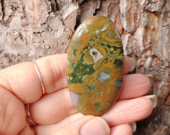 May include: An oval-shaped gemstone with a mix of colors, including green, brown, and white, is held in a hand. The stone has a polished surface and a unique pattern. The word "Front" is written in white at the bottom right.