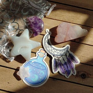 May include: A collection of crystals and stickers on a wooden surface. The crystals include a star, a purple sphere, and a pink cloud shape. Stickers feature a dreamcatcher, a potion bottle, and a star. A silver mesh bag is also present.