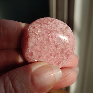 May include: A round, pink gemstone with a textured surface. The stone has a light pink color with a slightly rough texture. The gemstone is held in a hand, with the background out of focus.