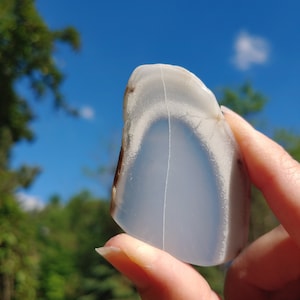 Blue chalcedony natural crystal slice. Small blue and white stone. Meditation stone pictured in hand. Bright sunlight, blue sky, green trees in background.