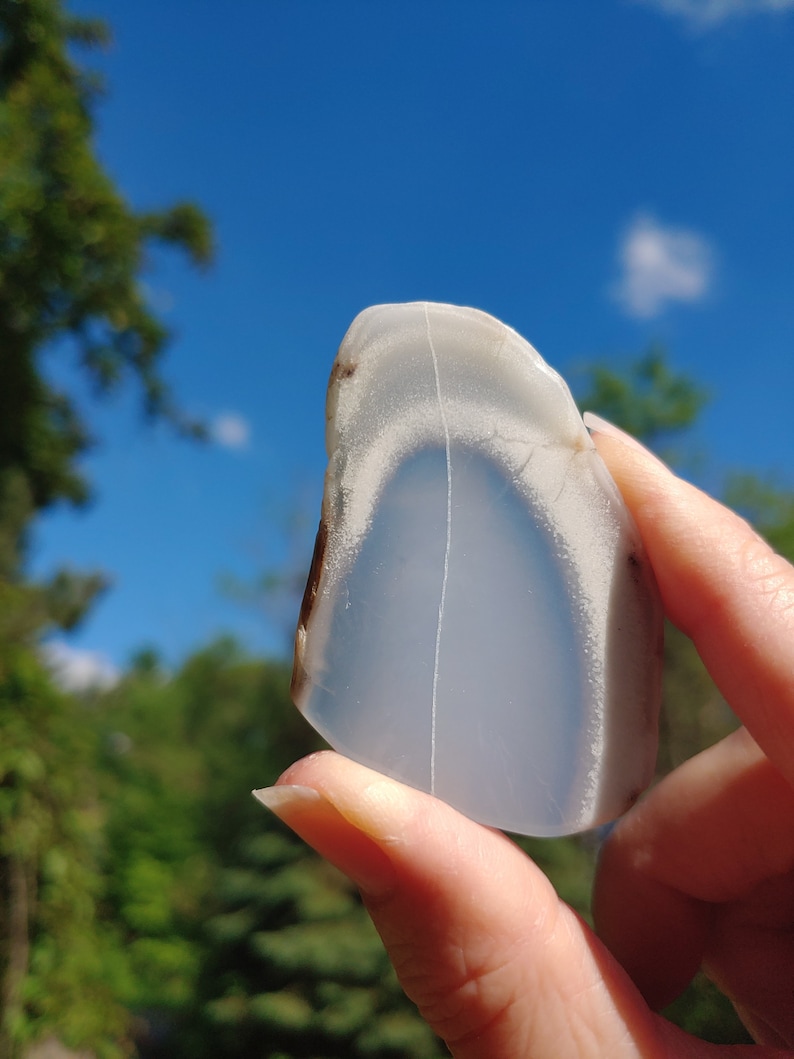 Blue chalcedony natural crystal slice. Small blue and white stone. Meditation stone pictured in hand. Bright sunlight, blue sky, green trees in background.