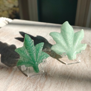 May include: Two green leaf-shaped decorative objects. One is a darker green, the other is a lighter green. Both are carved with leaf details and sit on clear plastic stands. The objects are on a light-colored wooden surface.