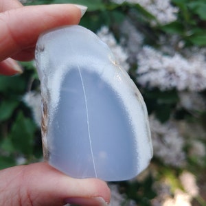 Blue chalcedony natural crystal slice. Small blue and white stone. Meditation stone pictured in hand. Shaded light, blooming lilac in background. Hand on the left side.
