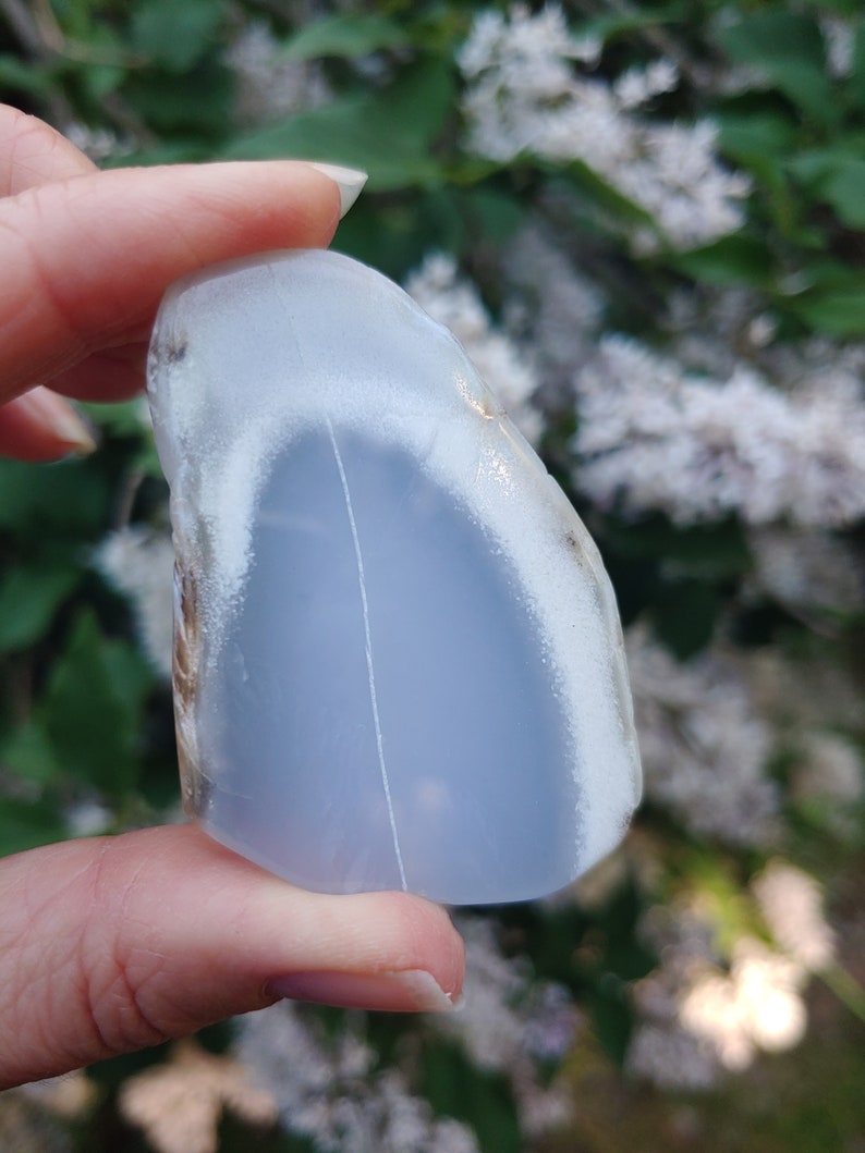 Blue chalcedony natural crystal slice. Small blue and white stone. Meditation stone pictured in hand. Shaded light, blooming lilac in background. Hand on the left side.