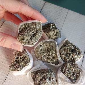 May include: A collection of pyrite specimens, also known as fool's gold, presented in individual white paper packaging. The metallic, golden-colored stones have a crystalline structure and are held by a hand against a light wood background.