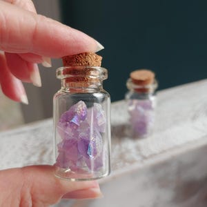 May include: A small, clear glass bottle filled with iridescent purple crystals, topped with a cork stopper. The crystals have a crystalline structure and reflect light. Another similar bottle is visible in the background.