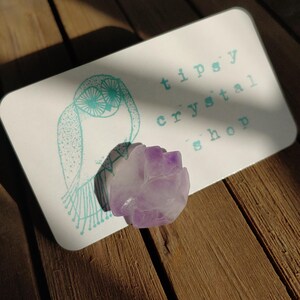 May include: A light purple crystal rose sits on a white card with the text "tipsy crystal shop" and a teal owl illustration. The card rests on a wooden surface, with sunlight casting shadows.