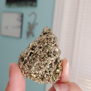 May include: A rough, golden-colored pyrite specimen, also known as fool's gold, is held up against a soft blue background. The mineral has a crystalline structure and a metallic luster, reflecting light. The pyrite is a natural, raw gemstone.