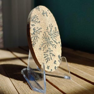 May include: A tan, oval-shaped stone with dark gray, fern-like patterns, displayed on a clear acrylic stand. The stone is positioned on a wooden surface, with a dark green background visible. The lighting highlights the texture and details of the stone.