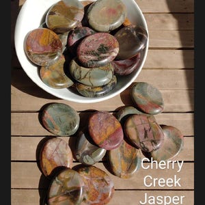 May include: A collection of Cherry Creek Jasper stones in a white bowl and scattered on a wooden surface. The stones display a variety of colors, including red, brown, green, and tan. The text "Cherry Creek Jasper" is visible in the image.