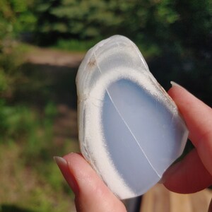 Blue chalcedony natural crystal slice. Small blue and white stone. Meditation stone pictured in hand. Bright sunlight, green trees in background.