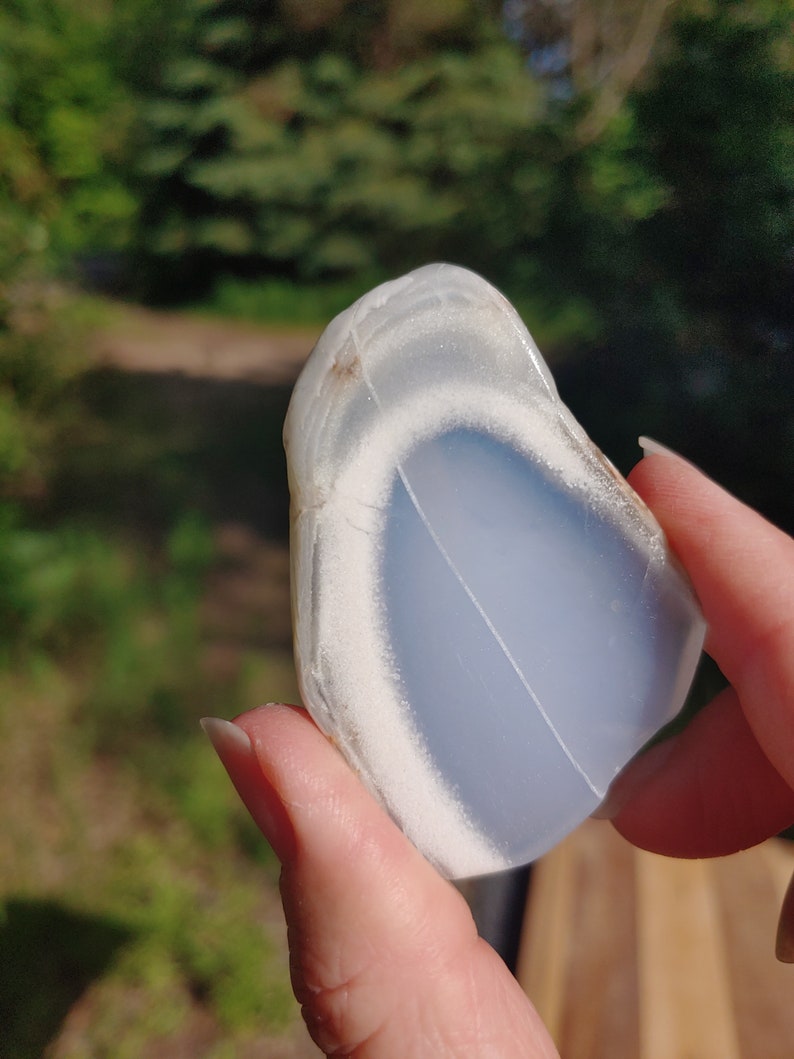 Blue chalcedony natural crystal slice. Small blue and white stone. Meditation stone pictured in hand. Bright sunlight, green trees in background.