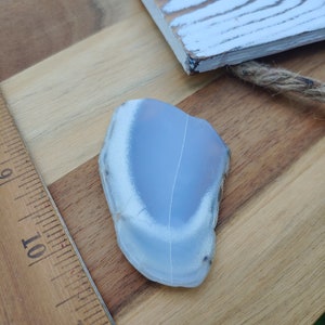 Blue chalcedony natural crystal slice. Small blue and white stone. Meditation stone pictured on butcher block wood table, next to vintage wood ruler. Shaded light, cool blue undertones.