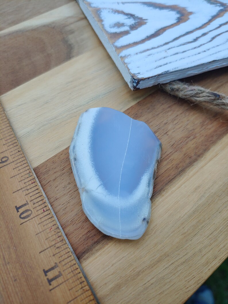 Blue chalcedony natural crystal slice. Small blue and white stone. Meditation stone pictured on butcher block wood table, next to vintage wood ruler. Shaded light, cool blue undertones.