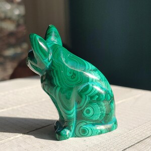 May include: A green malachite cat figurine, showcasing a sitting pose with its ears perked up. The stone's surface displays intricate patterns of varying shades of green, creating a unique, natural aesthetic. The figurine is a decorative item.
