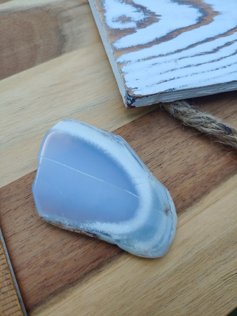 Blue chalcedony natural crystal slice. Small blue and white stone. Meditation stone pictured on butcher block wood table, next to vintage wood ruler. Shaded light, cool blue undertones.