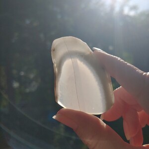 Blue chalcedony natural crystal slice. Small blue and white stone. Meditation stone pictured in hand. Bright backlight, blue sky, green trees in background.