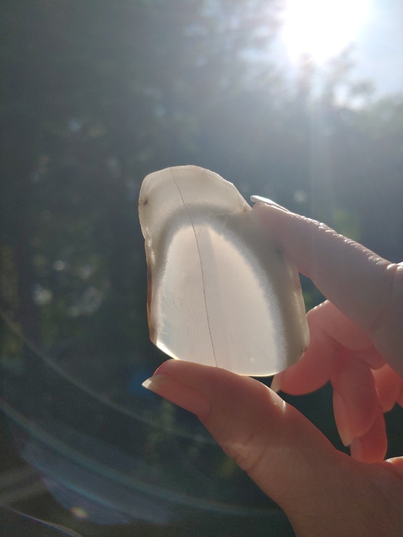 Blue chalcedony natural crystal slice. Small blue and white stone. Meditation stone pictured in hand. Bright backlight, blue sky, green trees in background.