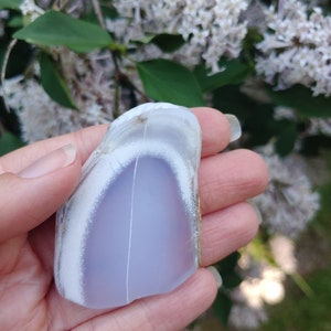 Blue chalcedony natural crystal slice. Small blue and white stone. Meditation stone pictured in hand. Shaded light, blooming lilac in background. Hand on the left side of frame.