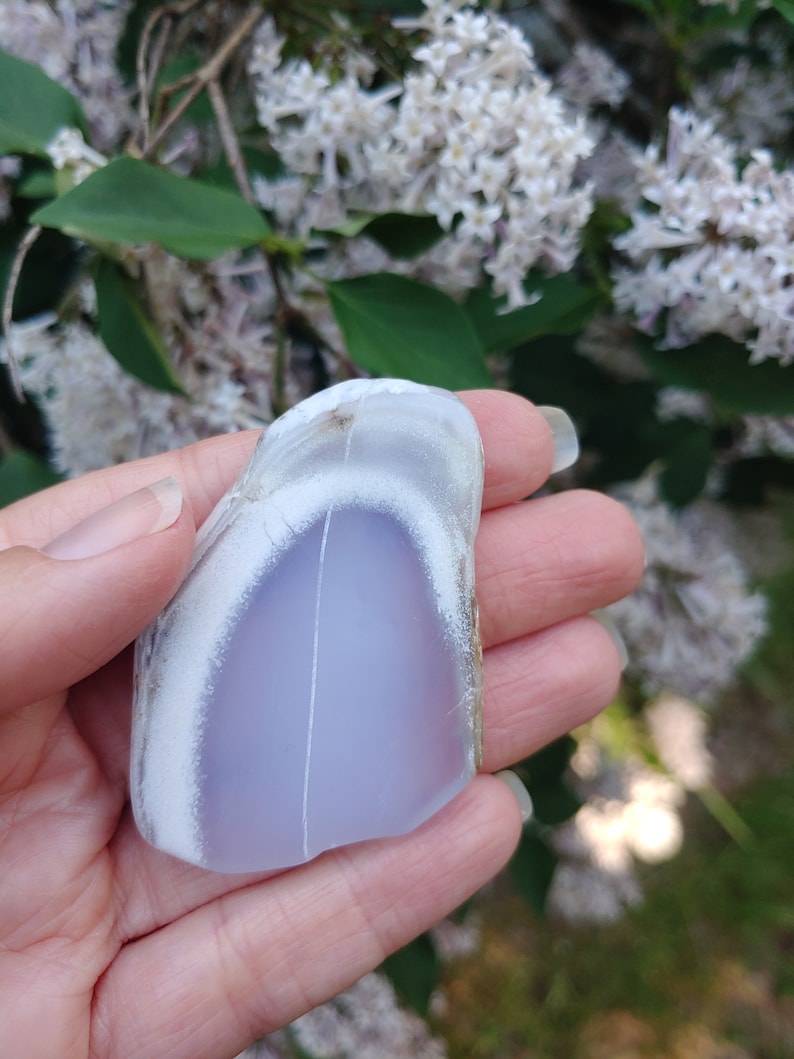 Blue chalcedony natural crystal slice. Small blue and white stone. Meditation stone pictured in hand. Shaded light, blooming lilac in background. Hand on the left side of frame.