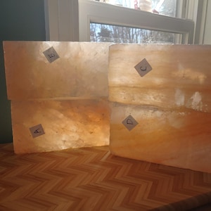 May include: Four rectangular, translucent, amber-colored salt blocks are stacked on a wooden surface. Each block has a small, white label with a letter. The blocks are illuminated by sunlight from a nearby window.