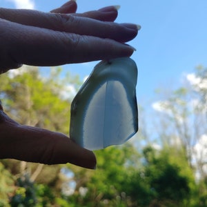 Blue chalcedony natural crystal slice. Small blue and white stone. Meditation stone pictured in hand. Bright backlight, blue sky, green trees in background.