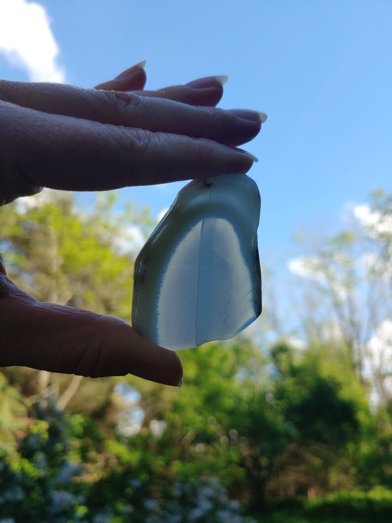 Blue chalcedony natural crystal slice. Small blue and white stone. Meditation stone pictured in hand. Bright backlight, blue sky, green trees in background.