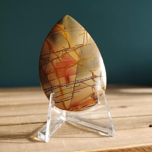 May include: A teardrop-shaped polished gemstone, showcasing a blend of beige, orange, and brown hues with intricate dark vein-like patterns. The stone is displayed on a clear acrylic stand, set against a teal backdrop.