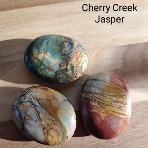 May include: Three polished Cherry Creek Jasper stones in shades of green, brown, red, and cream. The stones have an oval shape and are displayed on a wooden surface. The text "Cherry Creek Jasper" is visible.