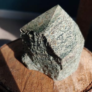 May include: A rough-hewn, green and gray stone crystal point with a flat top. The stone is set on a round, wooden base with visible wood grain. The crystal has a natural, textured surface.