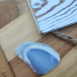 Blue chalcedony natural crystal slice. Small blue and white stone. Meditation stone pictured on butcher block wood table, next to vintage wood ruler. Shaded light, cool blue undertones.