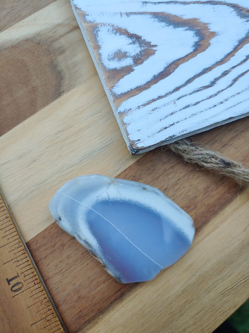 Blue chalcedony natural crystal slice. Small blue and white stone. Meditation stone pictured on butcher block wood table, next to vintage wood ruler. Shaded light, cool blue undertones.