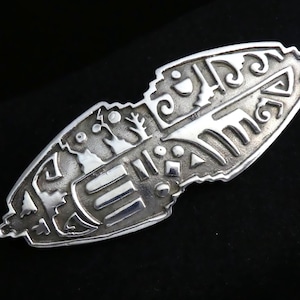 Puede incluir: Un broche de plata con un diseño intrincado de formas geométricas y árboles estilizados. El broche está hecho de una sola pieza de metal y tiene un acabado liso y pulido.