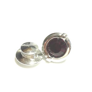 May include: A silver lapel pin featuring a deep red, circular gemstone. The pin has a classic design, with a round, domed head and a separate backing piece. The gemstone is set in a bezel setting, against a white background.