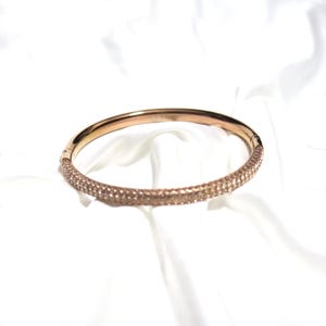 Op de afbeelding: Een roségouden armband met een band ingelegd met glinsterende kristallen. De armband ligt op een witte, zijdeachtige achtergrond. De kristallen bedekken de helft van de armband, wat een vleugje elegantie toevoegt.