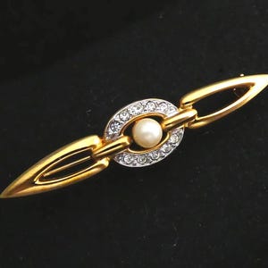 May include: Gold-toned brooch with a central oval design encrusted with clear crystals, surrounding a single pearl. The brooch has elongated, pointed ends and connecting links, creating a sophisticated, elegant accessory.