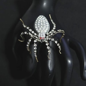 Puede incluir: Broche de araña plateada con detalles de cristal transparente y ojos rojos. El cuerpo de la araña está cubierto de cristales brillantes, y sus patas están adornadas con cristales más pequeños. Un accesorio único y llamativo.