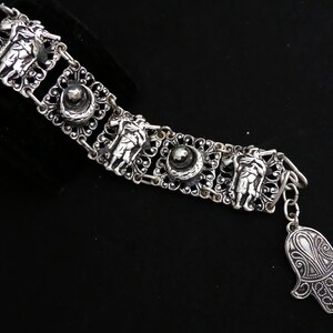 Può includere: Bracciale argentato con maglie in filigrana ornate con figure scolpite e dettagli circolari. Un ciondolo a forma di mano aggiunge un tocco unico. Il bracciale è esposto su sfondo nero.