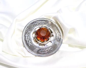 May include: A circular silver-toned brooch featuring an embossed thistle pattern around the perimeter and a large, faceted, amber-colored stone set in the center. The metal surface has a polished finish with intricate detailing.
