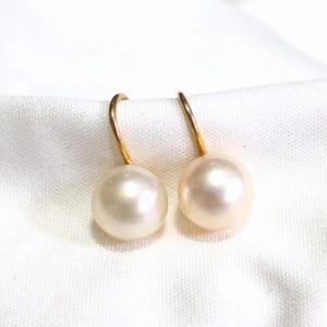 May include: A pair of drop earrings featuring round, cream-coloured pearls suspended from curved gold-tone metal hooks. The pearls have a smooth, lustrous finish and are positioned side by side, showcasing a simple and classic design aesthetic.
