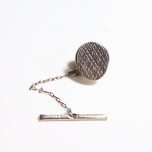 May include: A silver-toned tie tack featuring a circular head with a textured, cross-hatched pattern. The circular piece is connected by a delicate, thin metal chain to a slender, horizontal bar fastener designed to secure the accessory in place.