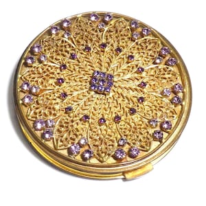 Könnte beinhalten: Ein runder, goldfarbener Kompaktpuder mit aufwendigen Filigranarbeiten. Der Puder ist mit zahlreichen kleinen, runden, lila Edelsteinen verziert. Das Design ist symmetrisch, mit einem zentralen Quadrat aus lila Steinen. Der Puder liegt auf weißem Hintergrund.