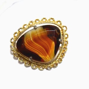 May include: A vintage brooch featuring a large, teardrop-shaped stone in brown and orange hues. The stone is set in a gold-toned metal frame with a decorative, scalloped edge. A classic piece of jewellery.