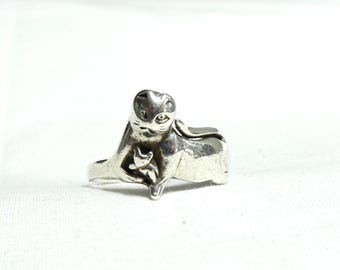May include: A silver-toned ring featuring a cat cradling a smaller kitten. The ring has a detailed design, with the cats' features clearly visible. The ring is set against a white background.