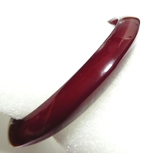 May include: A polished, deep red bangle bracelet. The curved surface reflects light, showing the rich colour. The bracelet is a simple, elegant accessory.