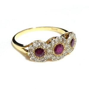 May include: A gold ring featuring three oval-cut rubies, each surrounded by a halo of small, clear gemstones. The ring's band is a simple, polished gold. The gemstones are set in a floral-like design.